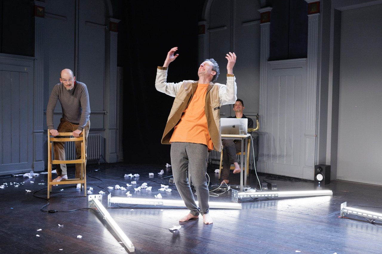 drei Schauspieler auf der Bühne, der Mann im Vordergrund wirft die Hände in die Luft.