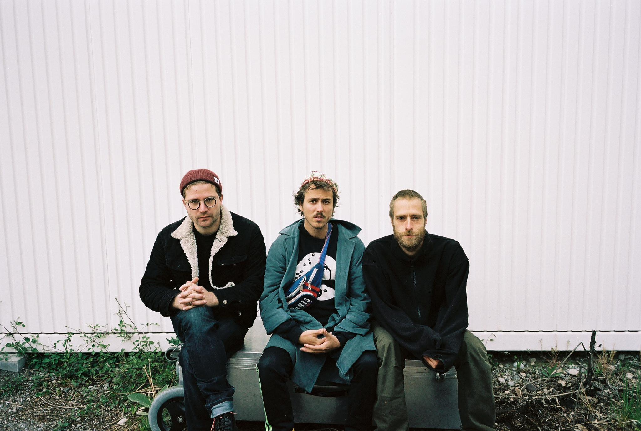 3 Menschen sitzen vor einem Container, schauen in die Kamera. Es ist ein Bandfoto