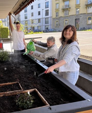 Man sieht Lukas, Marcel und mich (Nadine) am Hochbeet stehe, wie wir die Pflanzen tränken. Weil mir diese Arbeit sehr Freude macht, habe ich ein strahlendes Lächeln im Gesicht.