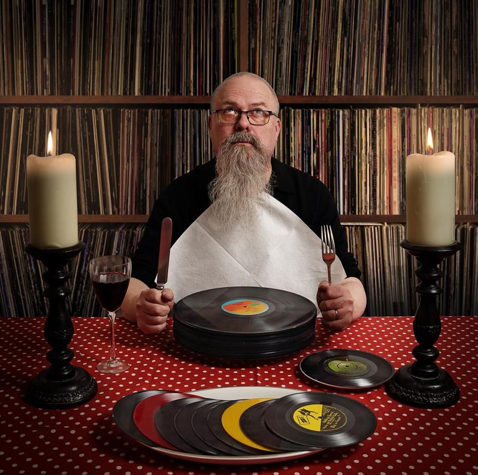 Ein mann sitzt an einem Tisch und hält Messer und GAbel in der Hand. Er trägt eine weisse Serviette. Der Mann hat einen grauen langen Bart und Glatze. Er schaut nach oben. Hinter ihm ist ein bildfüllendes Regal mit Schallplatten. Vor ihm liegt anstelle eines Tellers ein Haufen Platten.