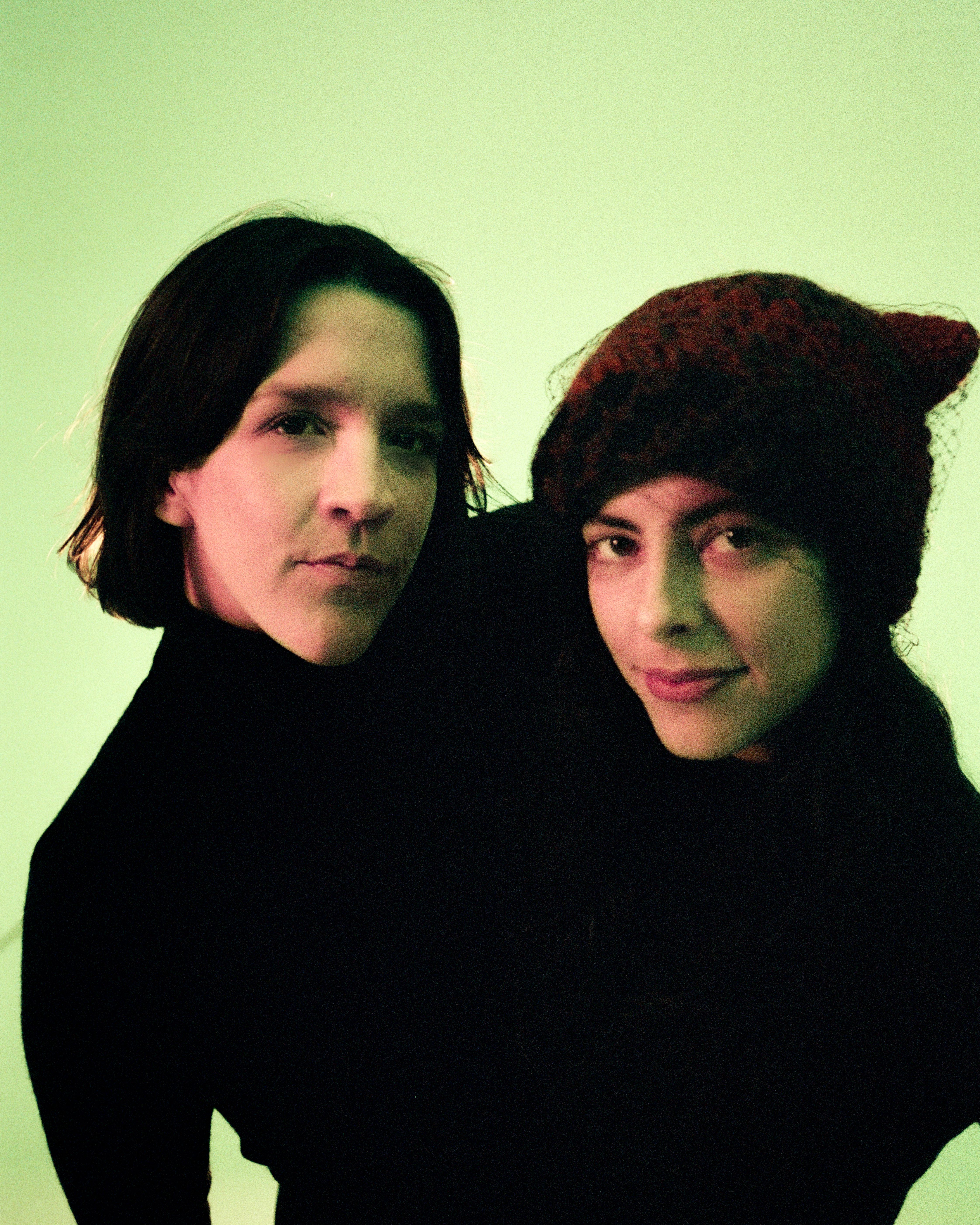 Zwie schwarzgekleidete weiblich gelesene Personen auf hellgrünem Grund schauen ind ei Kamera. Es ist nur ihr Oberkörper + Kopf zu sehen.