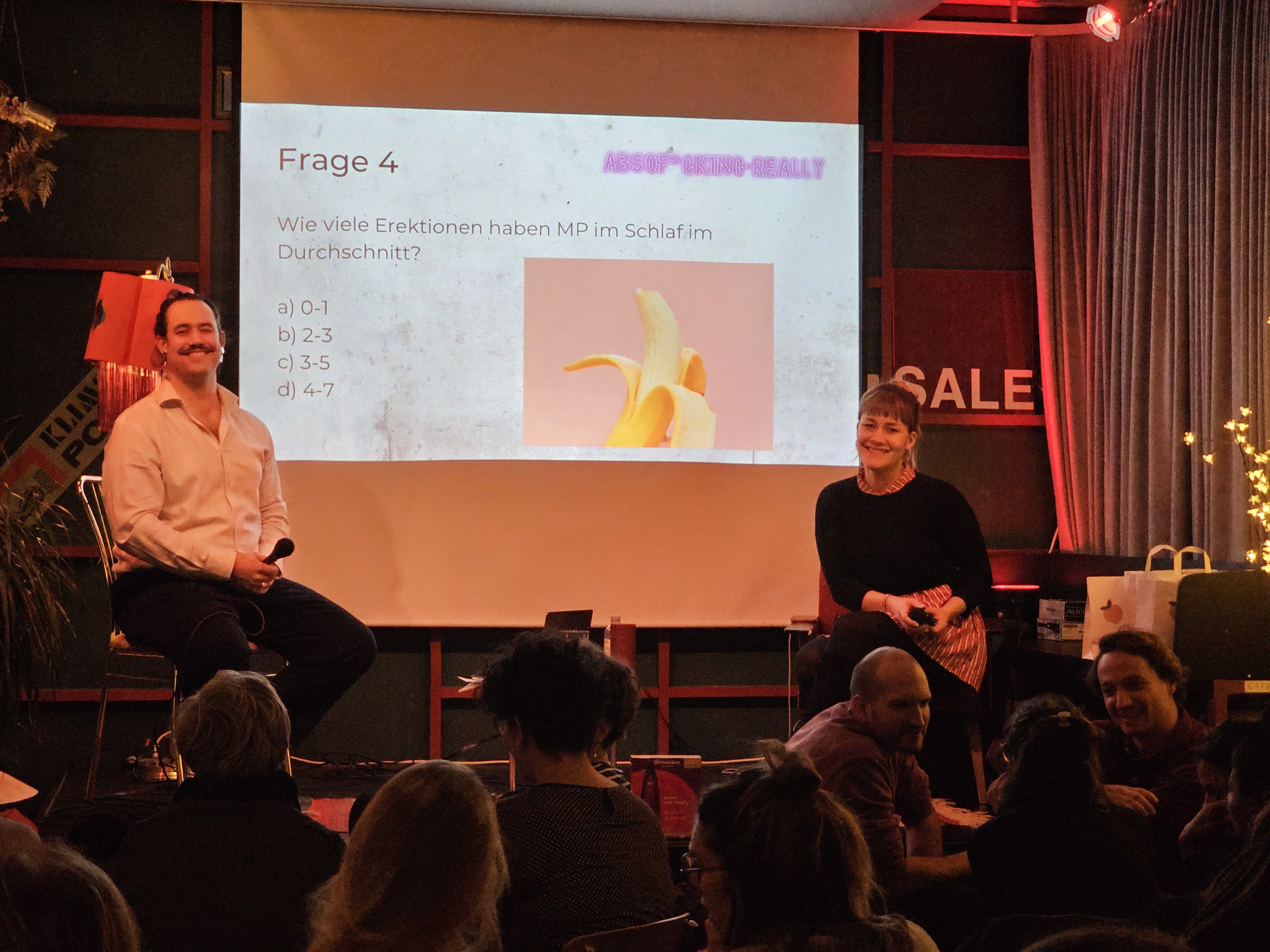Eine männliche Person und eine weibliche Person sitzen auf einer Bühne. Im Hintergrund ist eine Leindwand mit einer Projektion zu sehen.