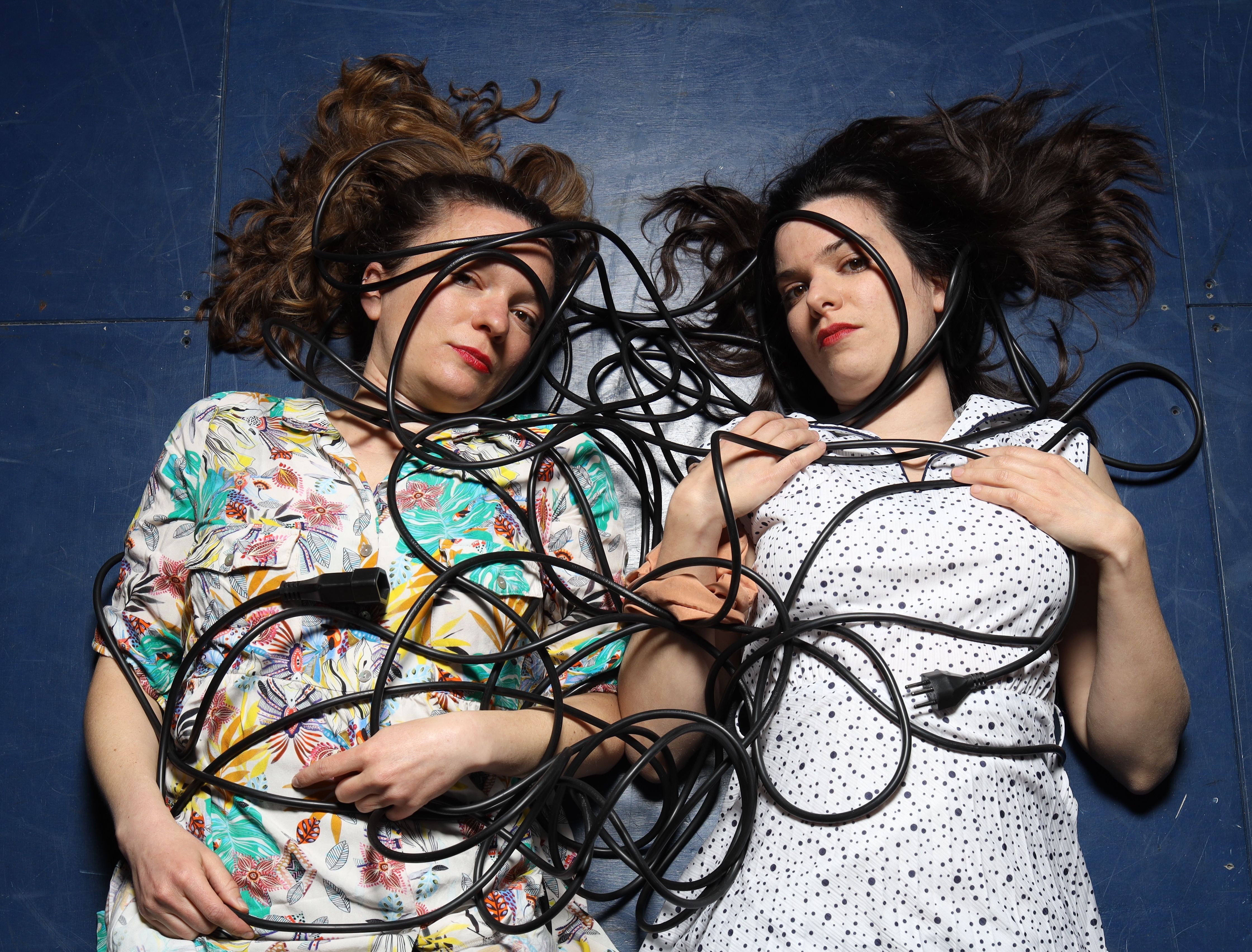 Zwei Frauen liegen auf einem blauen Boden. Auf ihnen drauf liegen schwarze Kabel.