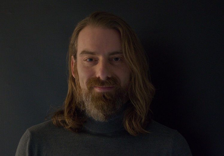 Ein Foto eines Mannes mit langem Bart und langen Haaren trägt einen dunklen Pullover. Das Foto zeigt ihn von der Brust aufwärts. Er steht vor einer dunklen Wand.
