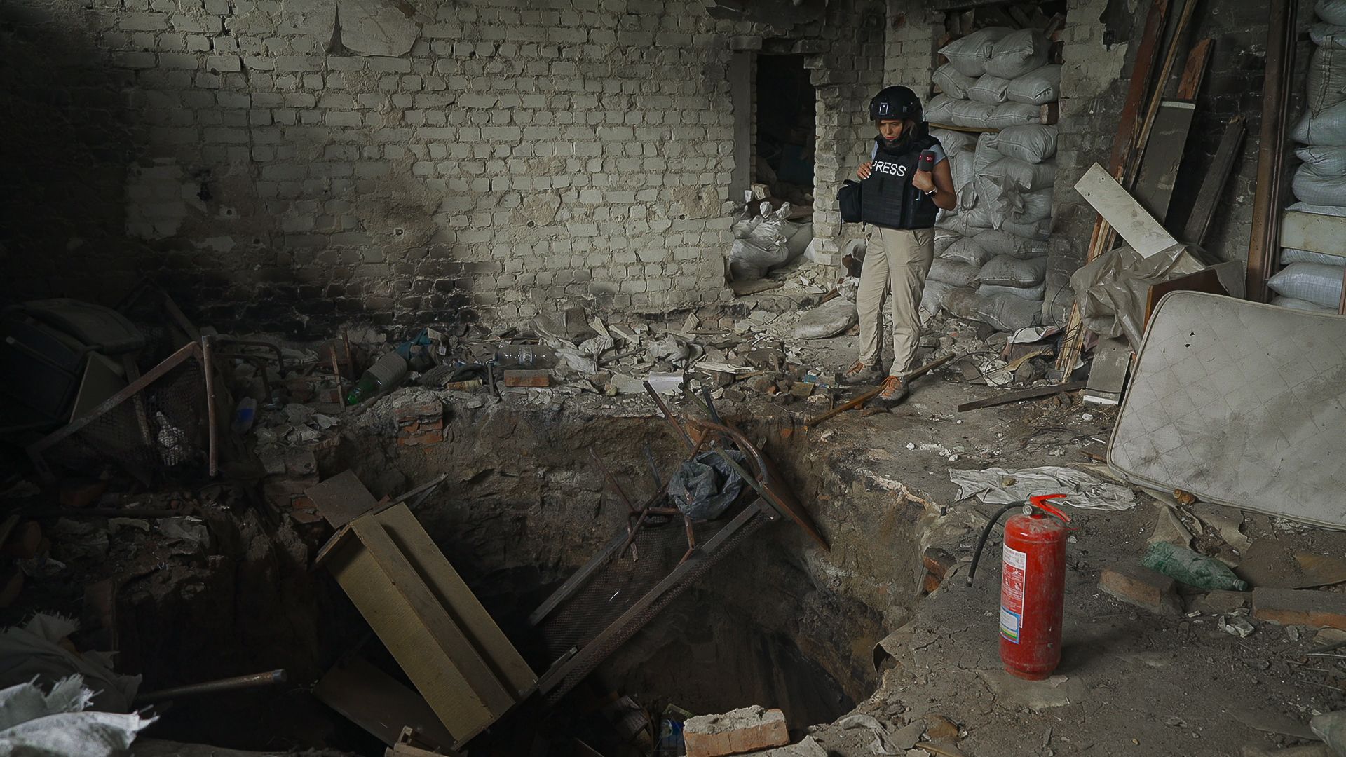 Ein Bild eines Bombenkraters in einem Haus. Rechts im Bild steht eine Reporterin in hellen Hosen und eine Schutzweste inkl. Helm.