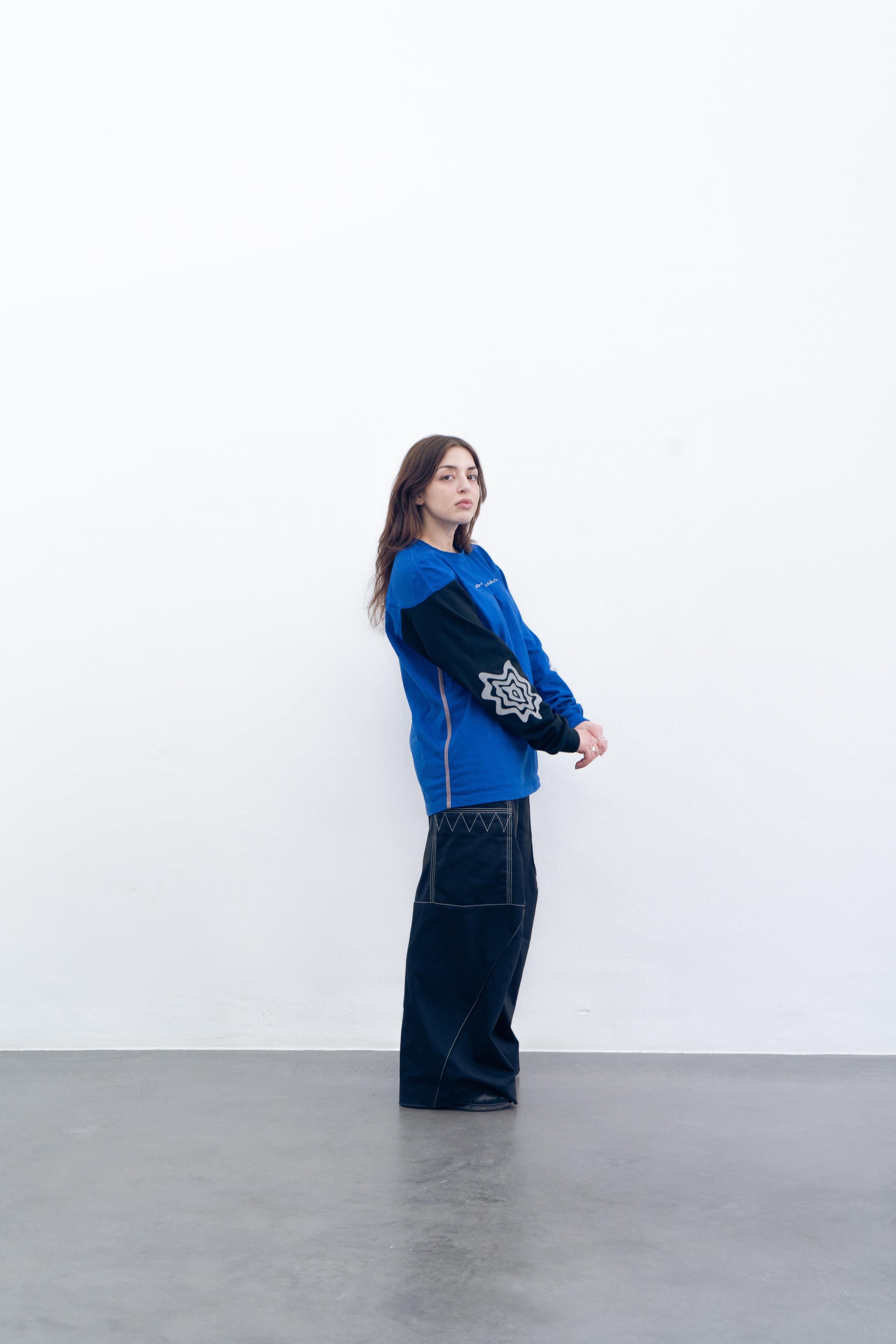 Eien Frau mit blauem übergrossem Shirt und schwarzen Hosen steht vor einer weissen Wand.