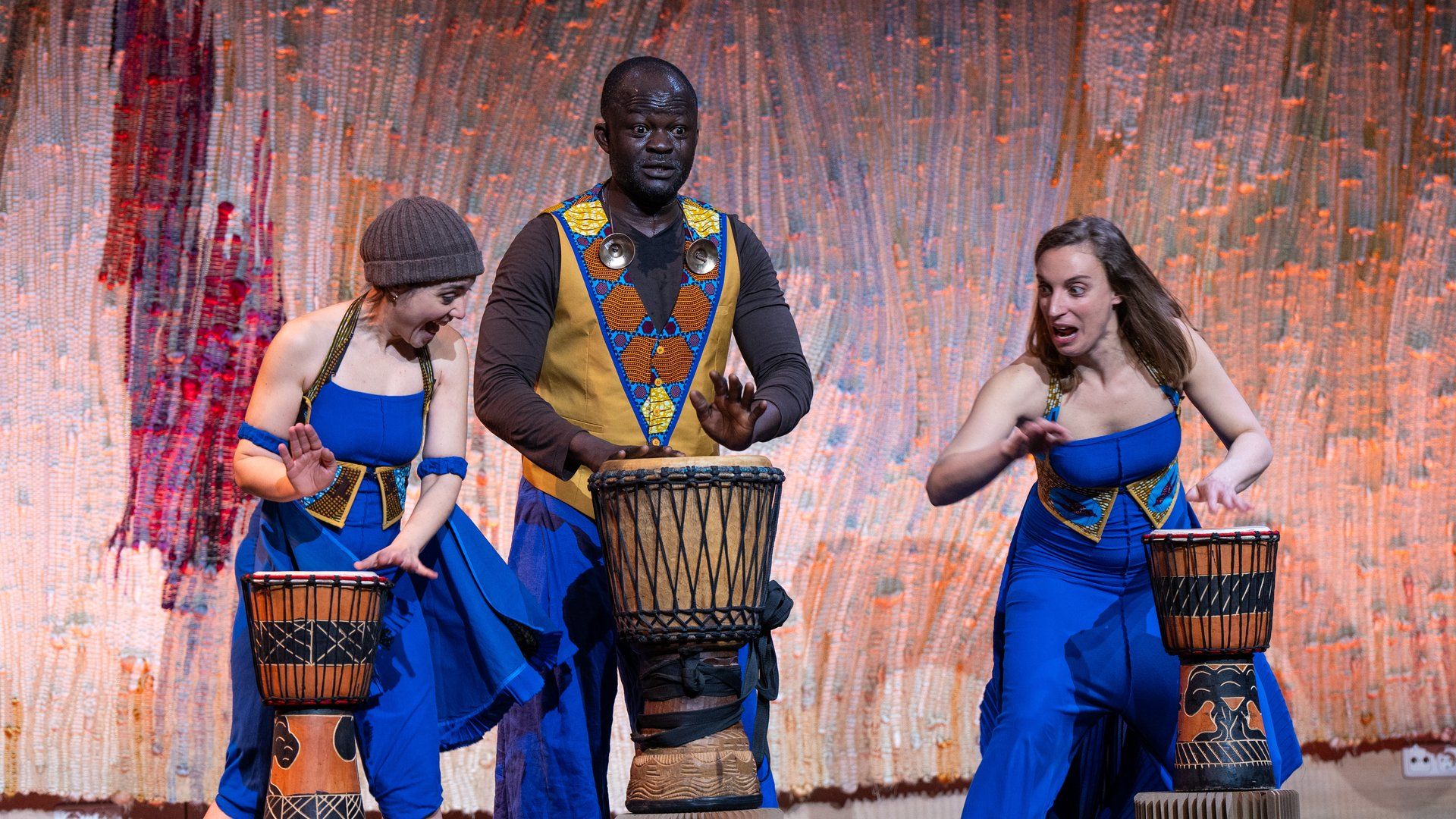 Drei Personen in farbigen Kleidern spielen jeweiles auf einer Trommel. Sie stehen vor einer rötlichen Wand mit Tüchern.