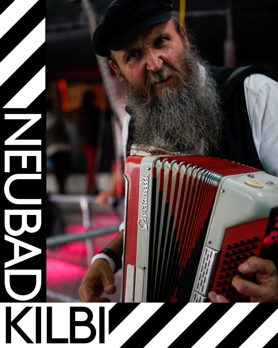 Ein bärtiger Mann mit schwarzer Schirmmütze und schwarzer Weste und einem Akkordeon. Am linken Rand steht "Neubad Kilbi"
