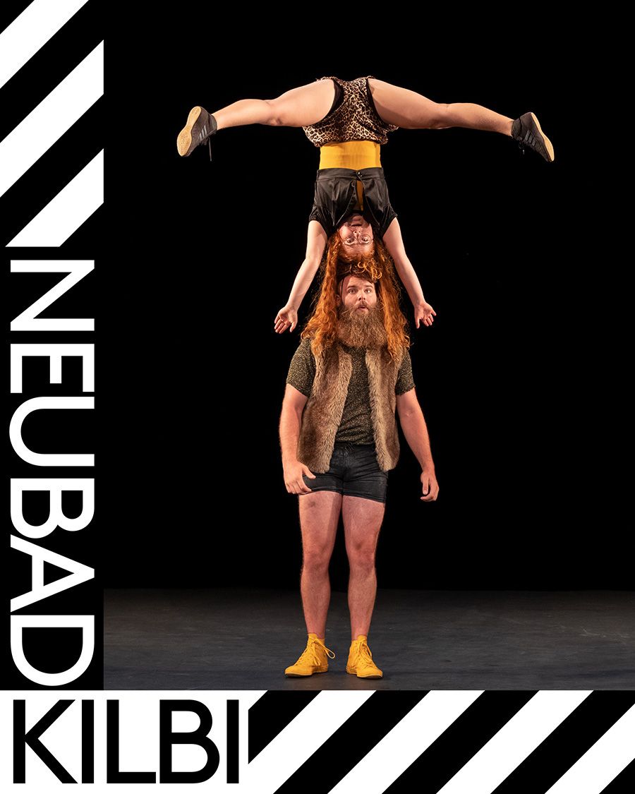 Ein grosser rotbärtiger Mann mit brauner Fellweste und gelben Schuhen balanciert eine rothaarige Frau mit schwarzen Klamotten und gelber Bauchbinde auf dem Kopf. Die Frau streckt ihre Beine horizontal aus und macht auf dem Kopf des Mannes einen Kopfstand. 