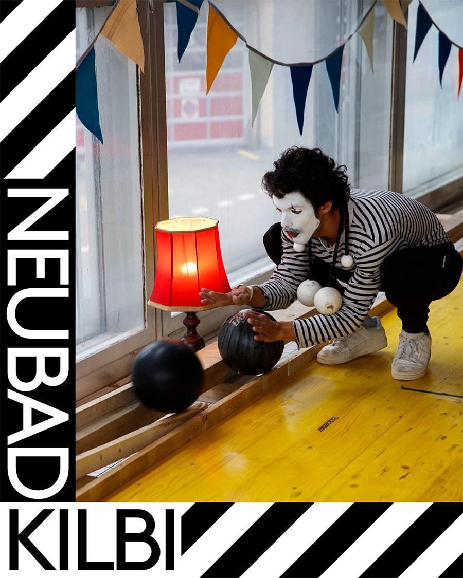 ein Pantomime spielt mit einer bowlingkugel an einer gelben bowlingbahn, neben der Bahn ist eine Tischlampe mit rotem Lampenschirm