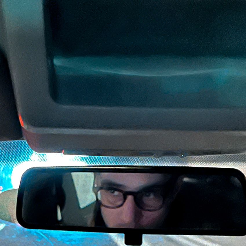 Foto von einem Autorückspiegel, in dem sich die die Augenpartie von einem bebrillten Mann spiegeln.