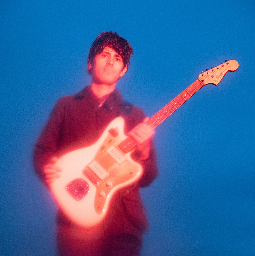 Ein Mann, der in rosa Licht beschienen wird und eine weisse Gitarre in der Hand hält vor einem blauen Hintergrund