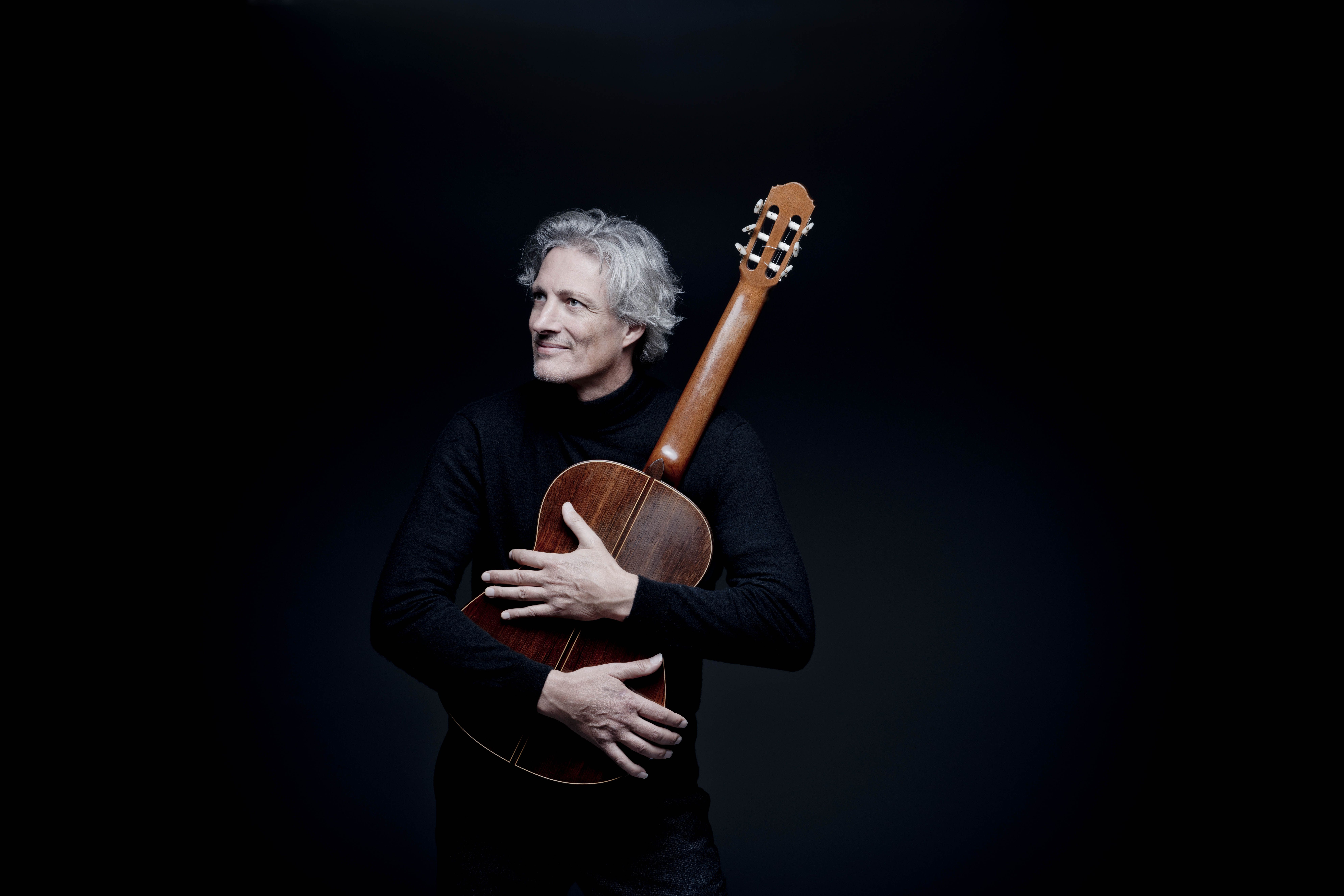 Ein Mann mit grauen Haaren, der eine Gitarre umarmt. Sein schwarzgekleideter Oberkörper verschmilzt mit dem schwarzen Hintergrund.