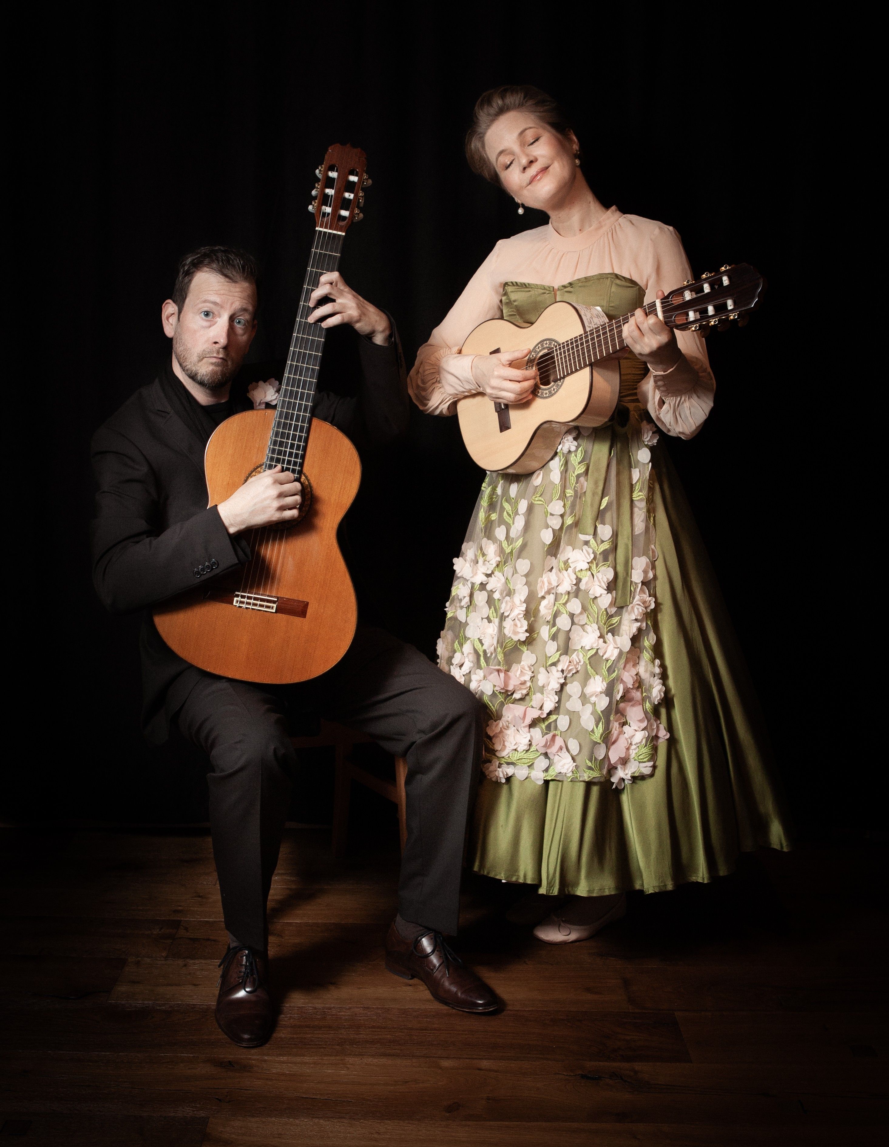 Ein Mann in Schwarz gekleidet und eine Frau in einem grünen Kleid. Beide halten eine Gitarre in der Hand.
