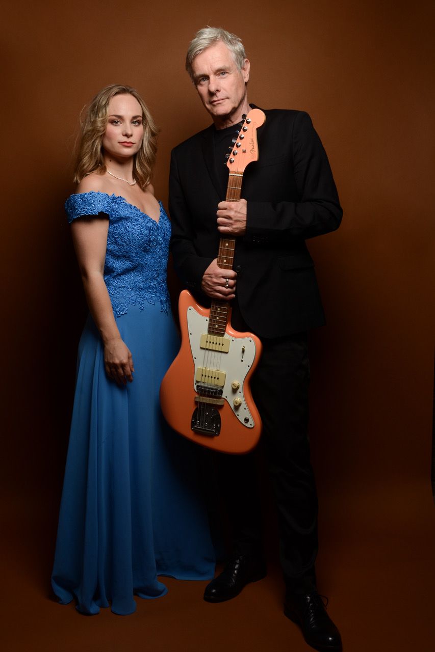 Eine Frau im blauem Kleid und ein schwarzgekleideter Mann mit Gitarre in der Hand vor einem dunkelbraunen Hintergrund