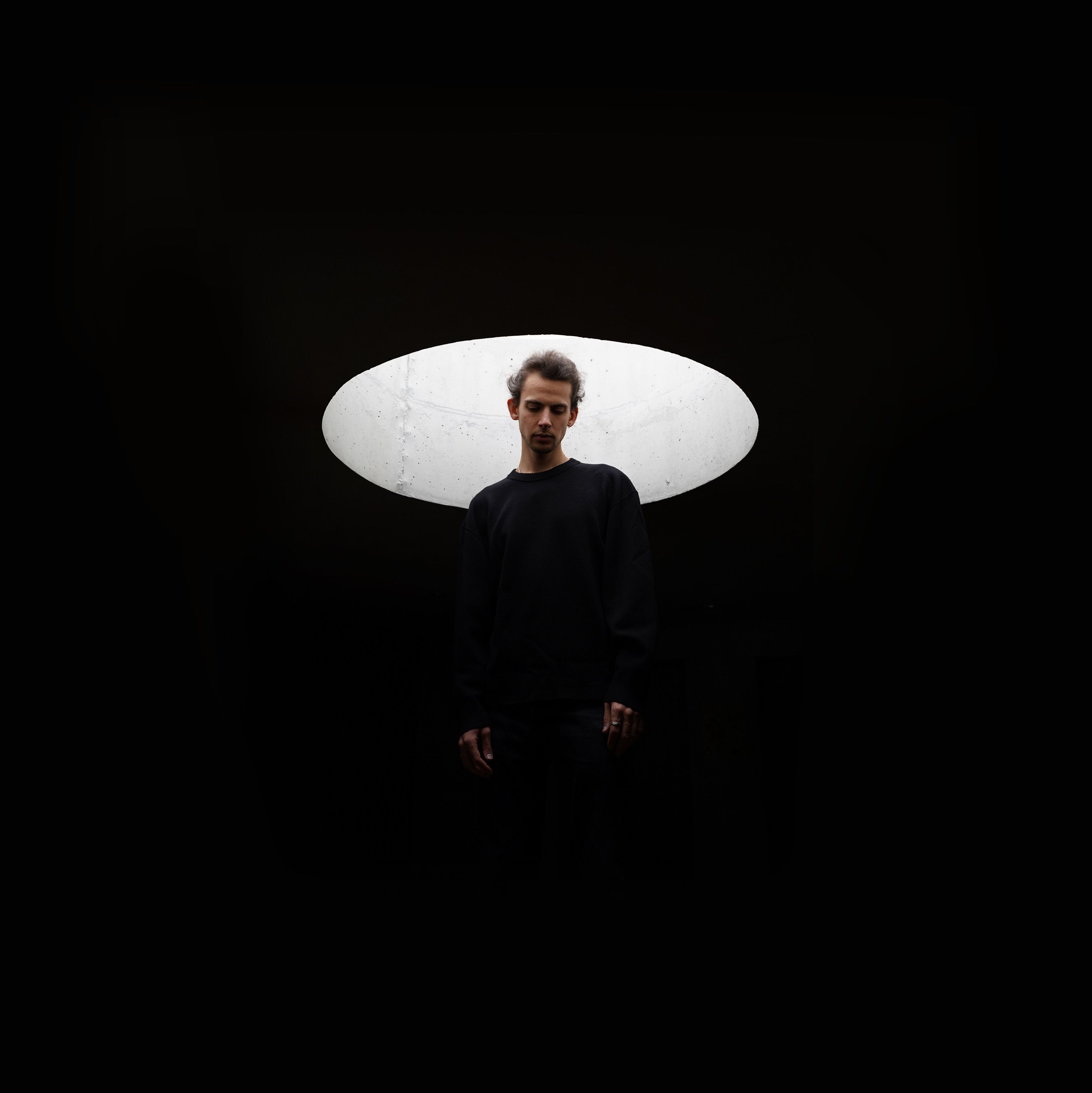 Eine Fotografie von einem schwarz gekleideten Mann auf schwarzem Hintergrund. Einzig die Arme und der Kopf sind vom Mann sichtbar. Hinter seinem Kopf befindet sich eine runde, hellgraue Öffnung, die ihn beleuchtet.  